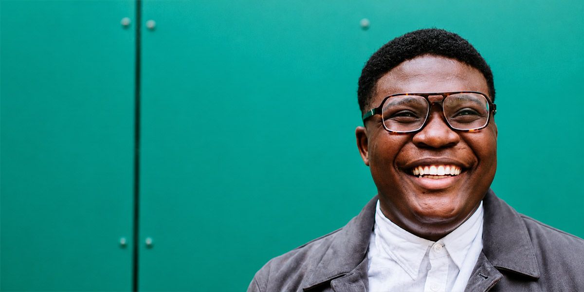 A person wearing glasses and similing, standing in front of teal wall